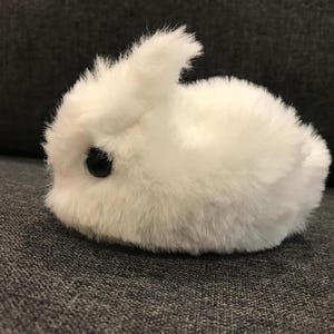 May include: A white fluffy bunny plush toy with black eyes. The bunny is lying on a gray fabric surface.