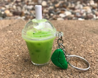 Könnte beinhalten: Ein Miniatur-Eisgetränk in Grün mit einem weißen Strohhalm und einem durchsichtigen Deckel. Ein grüner Blattanhänger ist an einem silbernen Schlüsselanhänger befestigt. Das Getränk befindet sich in einem durchsichtigen Plastikbecher.