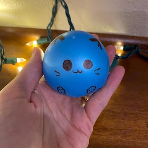 Pode incluir: Uma bola anti-stress azul com um rosto de desenho animado fofo desenhado nela. O rosto tem dois olhos, um nariz e uma boca.