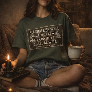 May include: A dark green t-shirt with the text "ALL SHALL BE WELL AND ALL SHALL BE WELL AND ALL MANNER OF THING SHALL BE WELL JULIAN OF NORWICH". The person is wearing denim shorts and holding a mug and a candle.