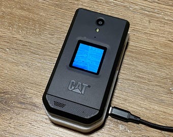May include: A black CAT branded flip phone with a square external display showing a blue screen, a camera lens, and a speaker grille. The device is connected to a black charging cable plugged into the side port on the right edge of the phone.
