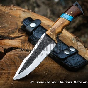 May include: A black-handled knife with a wooden handle and a blue inlay. The blade is made of steel and has a textured finish. The knife is resting on a piece of wood.
