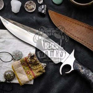 May include: A large, custom knife with a polished silver blade and a dark, patterned handle. The knife is displayed with a brown leather sheath, a small pouch, and various decorative stones. The blade features an etched design and the text "Black Mamba custom knives USA".