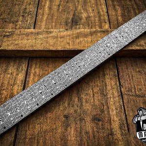 May include: A long, rectangular knife blade with a detailed, patterned design in shades of gray and black. The blade rests on a wooden surface. The Black Mamba custom knives logo is visible in the bottom right corner.