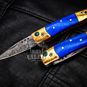 Puede incluir: Dos navajas plegables con diseños ornamentados. Los mangos presentan una sección central azul vibrante con detalles de puntos blancos, enmarcados por metal dorado. Las hojas tienen un aspecto texturizado y estampado. El texto "BLACK MAMBA custom knives USA" es visible.