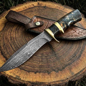 May include: A Damascus steel hunting knife with a dark wood handle and brass accents rests on a wooden surface. The knife is accompanied by a brown leather sheath. The Black Mamba logo is visible.