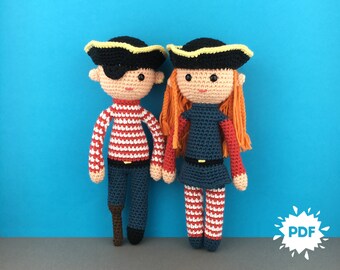 Könnte beinhalten: Zwei gehäkelte Piratenpuppen, eine mit einem rot-weiß gestreiften Shirt und einer blauen Hose, die andere mit einem blauen Kleid und rot-weiß gestreiften Leggings. Beide Puppen haben schwarze Hüte mit gelbem Besatz und orangefarbenes Haar.