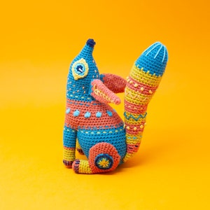 Puede incluir: Un juguete de animal de crochet con forma de zorro con un esquema de color azul, rosa, amarillo y verde. El zorro está sentado sobre una superficie amarilla.