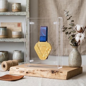 Puede incluir: Una medalla de oro con una cinta azul se exhibe en una caja de acrílico transparente. La medalla tiene el año 2021. La caja está montada sobre una base de madera. Un pequeño jarrón con algodón y vegetación está a la derecha.