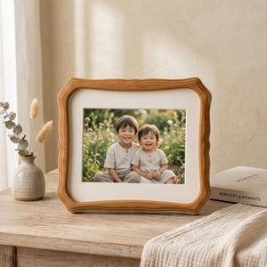 May include: A wooden picture frame with a light brown finish. The frame holds a photograph of two children smiling. The frame has a unique, wavy edge design. A small vase with dried flowers sits to the left of the frame.