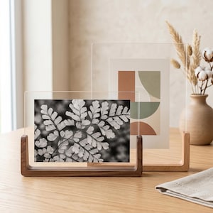 Puede incluir: Dos marcos de fotos flotantes de acrílico transparente con bases de madera. El marco delantero es horizontal con un estampado de hoja de helecho en blanco y negro. El marco trasero es vertical con un estampado geométrico en tonos terracota y verde salvia.
