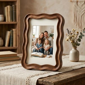 May include: A wooden picture frame with a wavy edge, displaying a family photograph. The frame is brown and rests on a wooden table with a beige runner. A vase of dried flowers is visible.