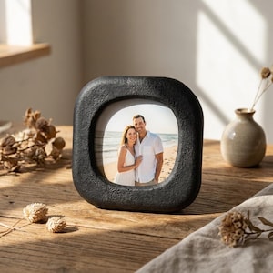 Puede incluir: Un marco de fotos negro, de forma cuadrada con esquinas redondeadas. El marco tiene una superficie texturizada y contiene una fotografía de una pareja en una playa. El marco está sobre una superficie de madera, con un pequeño jarrón y flores secas en el fondo.