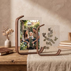 Könnte beinhalten: Zwei transparente Acryl-Fotorahmen mit gebogenen Halterungen aus Walnussholz. Ein Rahmen enthält ein Familienfoto, der andere einen botanischen Druck von Eukalyptusblättern. Die Rahmen stehen auf einem Holztisch mit einem neutralfarbenen Leinenläufer.