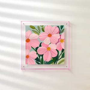 Peut inclure: Une impression d'art encadr&eacute;e repr&eacute;sentant des fleurs roses avec des centres orange et des feuilles vertes sur un fond bleu clair. L'&oelig;uvre est dans un cadre en acrylique transparent avec une bordure rose, mont&eacute;e sur un mur blanc.