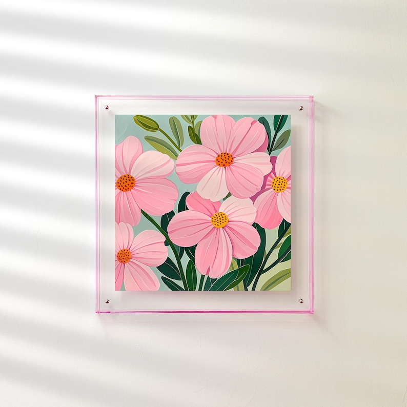 Peut inclure: Une impression d'art encadr&eacute;e repr&eacute;sentant des fleurs roses avec des centres orange et des feuilles vertes sur un fond bleu clair. L'&oelig;uvre est dans un cadre en acrylique transparent avec une bordure rose, mont&eacute;e sur un mur blanc.