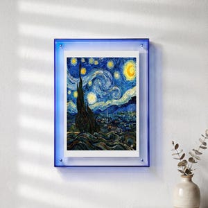 Peut inclure: Une impression encadr&eacute;e de l'&oelig;uvre "La Nuit &eacute;toil&eacute;e" de Van Gogh. L'impression est pr&eacute;sent&eacute;e dans un cadre en acrylique bleu, mont&eacute;e sur un mur blanc. L'&oelig;uvre pr&eacute;sente un ciel nocturne tourbillonnant avec des &eacute;toiles jaunes et un croissant de lune.