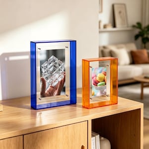 May include: Two acrylic photo frames, one blue and one orange, stand on a wooden surface. The blue frame holds an image of a clear geometric sculpture, while the orange frame displays colorful macarons. Both frames have a rectangular design.