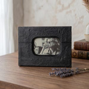 May include: A black rectangular picture frame with a textured surface. The frame holds a black and white photograph of a person wearing sunglasses. The frame is displayed on a wooden surface.