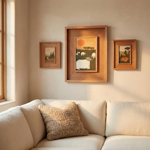 Op de afbeelding: Drie ingelijste landschapsschilderijen. Het grootste frame bevat een schilderij van een schaap in een veld met een ondergaande zon. De andere twee frames bevatten vergelijkbare landschappen, allemaal in houten frames. Een beige gebreid kussen ligt op een witte bank.