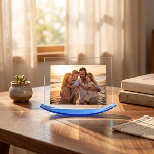 Puede incluir: Un marco de fotos de acrílico transparente con una base curva azul. El marco muestra una foto familiar de una madre, un padre y dos hijos en una playa. A la izquierda hay una pequeña planta suculenta en una maceta de cerámica.
