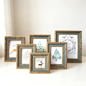 May include: A collection of six rectangular picture frames with ornate, distressed gold and blue-grey finishes. Each frame contains a unique holiday-themed illustration, including a reindeer, a Christmas tree, and text reading Joy to the World.
