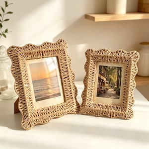 May include: Two rectangular picture frames with wavy, scalloped edges made from woven light tan natural fibre. The left frame displays a sunset over the ocean, while the right frame shows a tree-lined street scene with a building and outdoor seating.