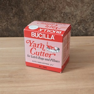 May include: A red and pink cardboard box for a Bucilla yarn cutter for latch rugs and pillows. The box features white text reading Yarn Cutter for Latch Rugs and Pillows, Cuts Yarn Approx. 2 1/2 inches, and Art. 7006, with a graphic illustration.