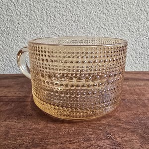 May include: A clear, amber-colored glass mug with a handle. The mug features a textured, bubble-like pattern across its surface. The mug is sitting on a wooden surface.