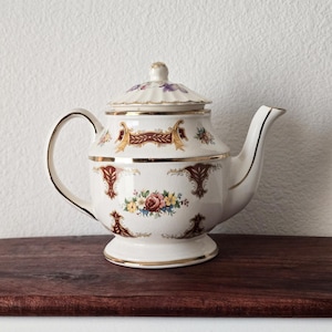 May include: A cream-colored ceramic teapot featuring a lid with a small knob, a curved handle, and a spout. The body is decorated with gold-trimmed maroon scrollwork patterns and a central floral arrangement of roses and small yellow flowers.