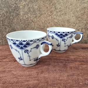 May include: Two white porcelain teacups with blue floral patterns and handles. The rims are decorated with a blue scalloped design. The cups are placed on a wooden surface against a neutral background.