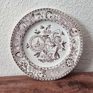 May include: A round, antique-style plate with a brown and white design. The plate features a central image of a sailboat, surrounded by floral and geometric patterns. The rim has a detailed border with various decorative elements.