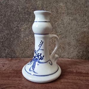 May include: A white ceramic candlestick holder with a blue floral design. The holder has a bulbous base, a cylindrical body, and a handle. Blue stripes accent the top and bottom.