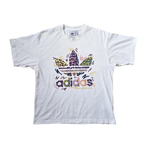 May include: White t-shirt featuring a colorful Adidas logo with a unique, abstract design. The logo incorporates various patterns and colors, including yellow, pink, blue, and black. The shirt has short sleeves and a crew neck.