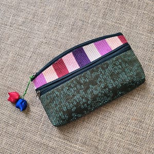 May include: A rectangular, dark green zippered pouch with a floral pattern and a striped top in shades of pink, purple, and red. A small tassel with red and blue accents hangs from the zipper.