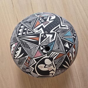 May include: A round, black ceramic pot with a geometric design and a frog illustration. The pot features a black and white geometric pattern with accents of pink, blue, and brown. A hole is visible near the top.