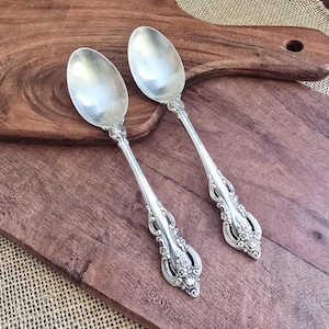May include: Two ornate silver spoons with detailed floral and scrollwork designs. The spoons are placed on a dark wooden cutting board. The spoons have a shiny, reflective surface.