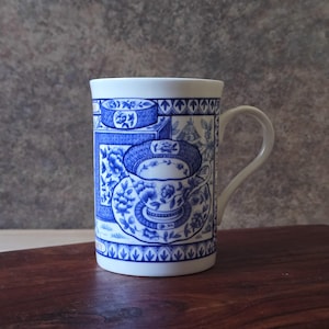 May include: A white ceramic mug featuring a blue and white transferware pattern. The design depicts a teacup and saucer set surrounded by floral motifs and geometric borders. The mug has a curved handle and a smooth, cylindrical body shape.
