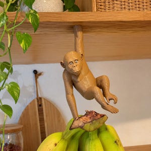 Puede incluir: Una figura de mono de color marrón claro impresa en 3D que cuelga de un estante de madera por un brazo, colocada directamente sobre un racimo de plátanos amarillos. El mono tiene un acabado liso y mate, rasgos detallados y ojos blancos.