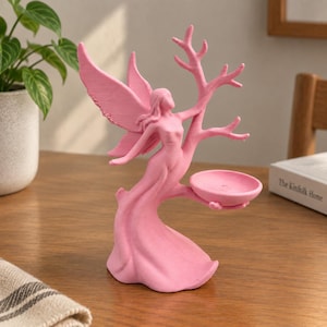 May include: A matte pink figurine depicting a winged figure standing beside a branching tree structure. The figure has long hair and a flowing gown that forms the base. A small, circular, bowl-shaped tray extends from the tree branches.