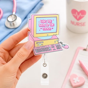 Può includere: Un porta badge con un'illustrazione di un computer retrò in rosa e giallo pastello. Lo schermo mostra il testo "take me back to the 2000s" con piccole icone a forma di cuore. Il design include una tastiera e un mouse collegato da un cavo.