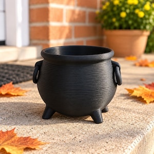 May include: A matte black cauldron-shaped planter with a rounded body, a wide rim, and three short legs. The exterior features a fine, horizontal ribbed texture and two small ring-shaped handles attached to the sides of the vessel.