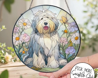 May include: Round stained glass suncatcher featuring a seated Old English Sheepdog with a light blue and cream coat, surrounded by daisies and other flowers. The dog's name, "Coca", is written at the bottom. Includes a hanging chain.