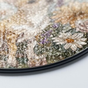 May include: A round decorative coaster with a floral design. The coaster features a detailed illustration of various flowers in shades of white, yellow, purple, and green. The coaster has a black border and a textured surface.