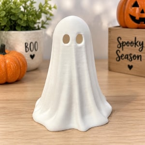 May include: A white, three-dimensional ghost figurine with a smooth, draped fabric-like texture. The ghost features two oval-shaped eye cutouts near the top of its rounded head and a wide, flared base that rests flat on a surface.