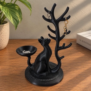 May include: A black jewelry stand featuring a cat sitting on a circular base. The cat's tail extends into a small bowl for rings, while a tall, branch-like structure rises behind the cat to hold necklaces and bracelets. The stand is matte black.