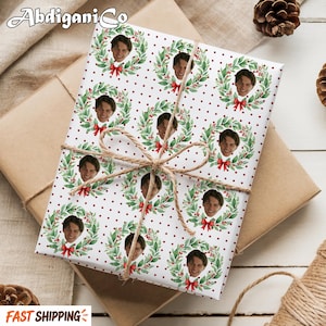 May include: White wrapping paper with a repeating pattern of green Christmas wreaths, red berries, and small red dots. Each wreath frames a photograph of a person's face and features a red bow at the bottom. The paper is tied with brown twine.