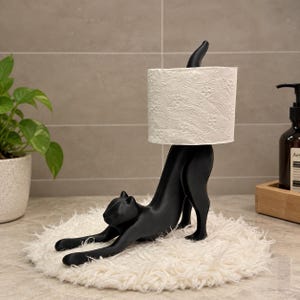 May include: A matte black cat-shaped toilet paper holder featuring a feline in a stretching pose. The cat's upright tail serves as a spindle for a roll of white toilet paper with a textured floral pattern, positioned on a round white faux fur mat.