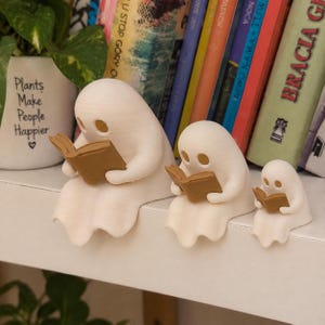 May include: Three off-white ghost figurines of varying sizes, each sitting on a ledge and holding a small brown book. The ghosts have smooth, rounded shapes with hollow eye sockets. A white planter with the text Plants Make People Happier is visible.