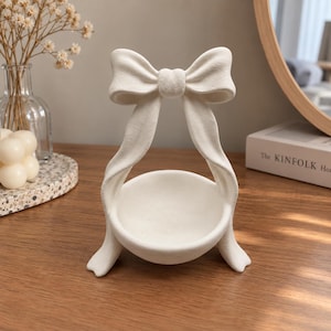 May include: A matte white ceramic trinket dish featuring a sculptural design with a large bow at the top. The bowl is circular and supported by two curved, ribbon-like legs that extend from the bow, creating a unified, solid white structure.